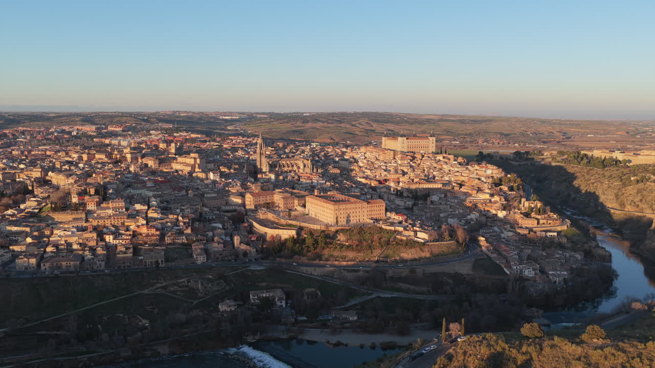 Tasainen ilmakuva Toledosta, Espanjasta, auringonlaskun aikaan. Auringonsäteet luovat kultaista valoa muinaisten rakennusten ylle, yllä on kirkas taivas ja alla näkyy Tagus-joen virta.