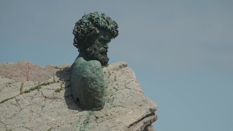 Ampia inquadratura del monumento a busto di Philippe Cousteau integrato nelle scogliere rocciose presso il Museo dell'Ancora a Salinas, Asturie, Spagna. Presenta onde oceaniche che si infrangono e una piattaforma panoramica elevata.