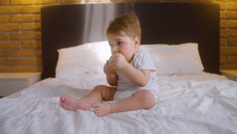 front view of a baby sitting on the bed in bedroom while bitting a green apple 1