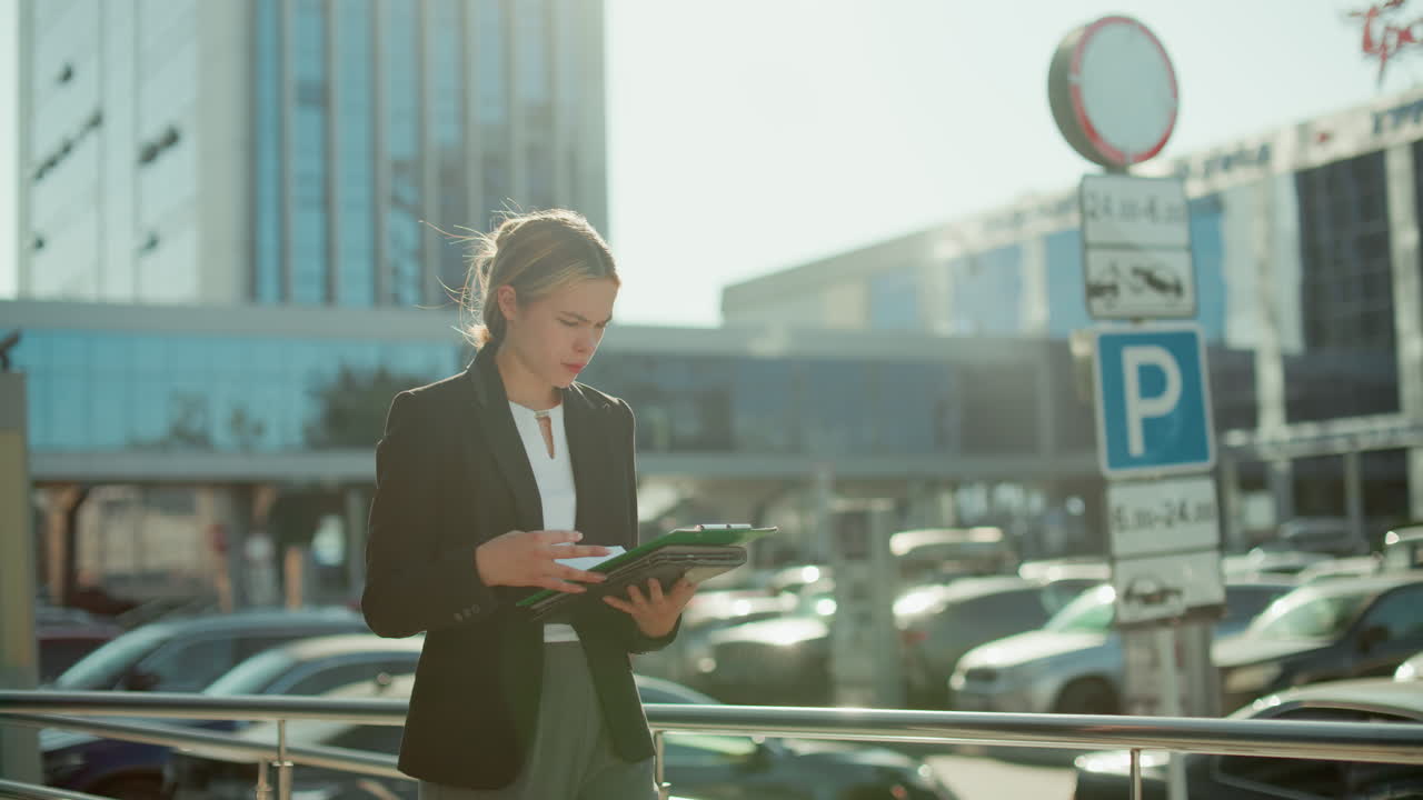 Bank manager in business suit walking outside urban office area reviewing documents in folder under bright sunlight with parked cars, street signs, and glass buildings in background