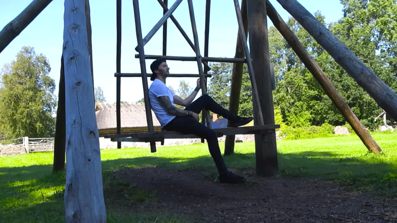 Man with tattoos, white shirt and a baseball cap swinging and sitting on a traditional wooden northern european swing during summer sunny day at an open air museum in Tallinn, straw roof houses also