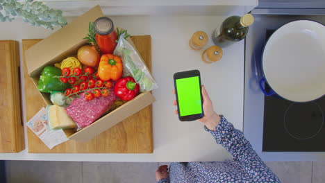 Fotografía-Cenital-De-Una-Mujer-En-La-Cocina-Con-Ingredientes-Frescos-Buscando-Una-Receta-En-Línea-En-Un-Teléfono-Móvil-Con-Pantalla-Verde,-Filmada-En-Cámara-Lenta