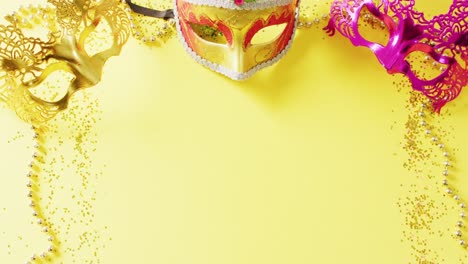 Video-of-three-masquerade-masks,-mardi-gras-beads-and-confetti-on-yellow-background-with-copy-space