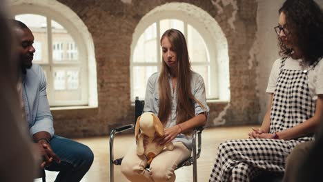 una chica rubia con cabello largo habla de su problema mientras sostiene un juguete blando en sus manos, y el resto de los participantes de la terapia de grupo apoyan a la chica y ponen sus manos en sus hombros