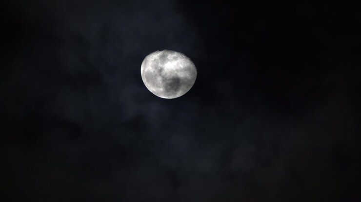 Crateras e cristas de uma lua branca e brilhante elevam-se no céu enquanto as nuvens passam por baixo à noite