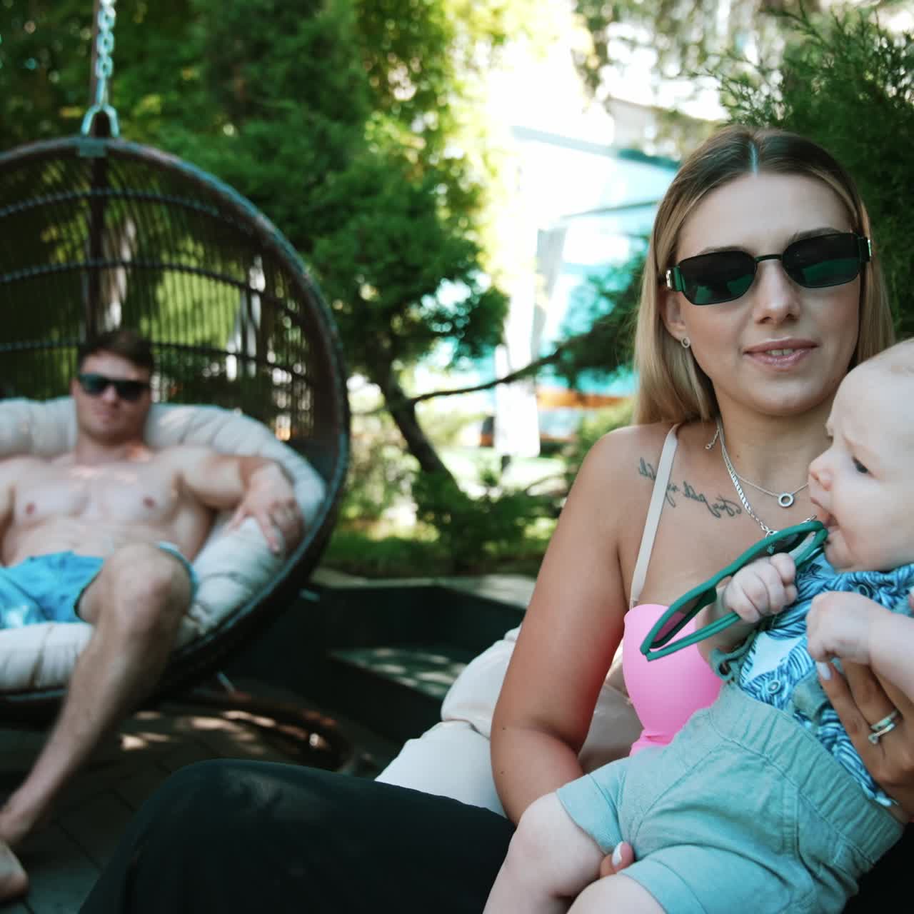 Kaukasisk familie med nyfødt, der slapper af sammen udendørs om sommeren. Kvinde holder baby, og mand solbader i baggrunden