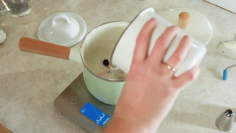 close up of hand pouring sugar and berries into white pot with wooden handle placed on digital kitchen scale for weight measurement during cooking preparation
