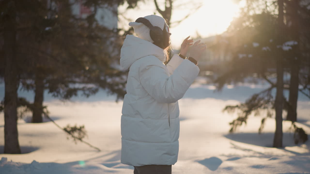 Side view of female figure donning sunglasses under bright winter sky while smiling joyfully breeze tousles hair light glints on lenses headphones rest on neck wearing puffer coat and beanie