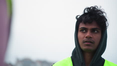 Young male worker wearing dark hoodie and neon reflective vest standing outdoors against bright overcast sky, gazing into distance with serious determined expression and readiness for work tasks
