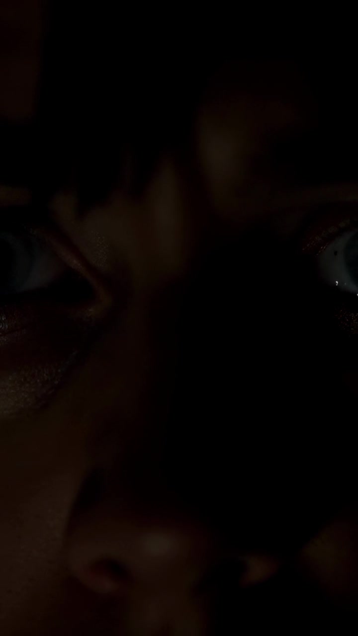 Vídeo vertical: mulher olhando fixamente com os olhos bem abertos enquanto a câmera do smartphone é ativada em uma sala mal iluminada