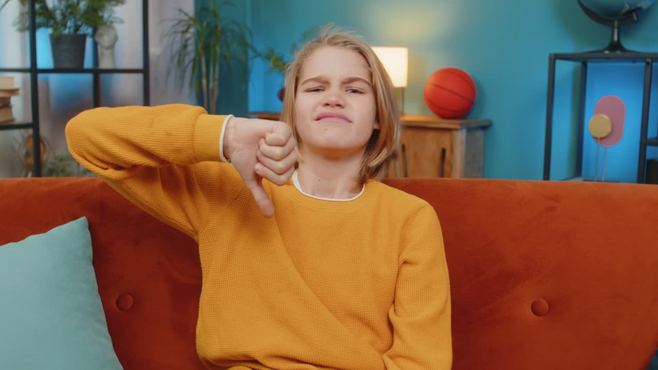 Teenager boy giving thumbs down frowning showing disappointment sitting at home dissatisfied