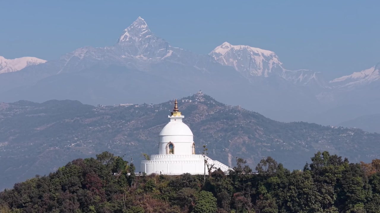 Ripresa aerea del monumento buddista bianco Shanti Stupa su una collina con imponenti cime himalayane innevate sullo sfondo.