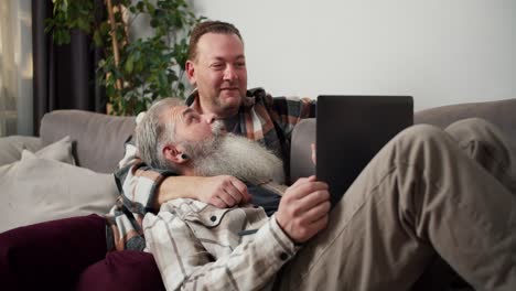 Un-Hombre-Feliz-De-Mediana-Edad-Con-Cabello-Gris-Y-Una-Exuberante-Barba-Gris-Con-Una-Camisa-A-Cuadros-Se-Acuesta-En-El-Regazo-De-Su-Novio-Moreno-Con-Una-Camisa-A-Cuadros-Y-Sostiene-Una-Computadora-Portátil-Gris-En-Sus-Brazos-Durante-Su-Pasatiempo-Común-Cerca-De-La-Computadora-Portátil-En-El-Sofá-Del-Apartamento.