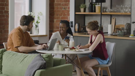 three  rommates having breakfast, using the laptop and taking notes while sitting together at kitchen table at home