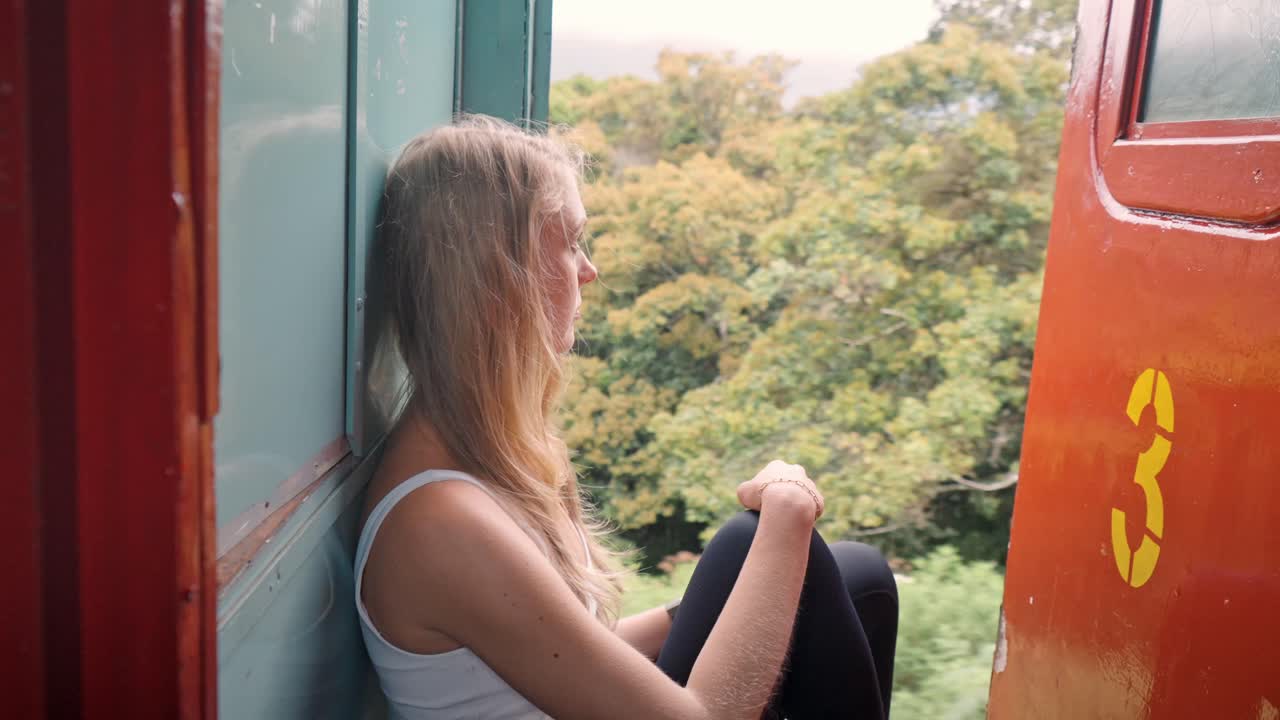 A woman sits peacefully on the open door of a moving train along the iconic Ella to Kandy route in Sri Lanka, taking in the breathtaking views of green hills, tea plantations, and dense jungle.