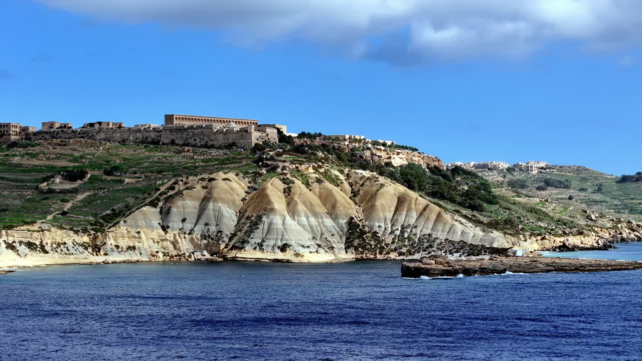 Fort Chambray in cima a suggestive scogliere argillose, che domina le acque blu intenso del Mediterraneo sotto cieli soleggiati durante una vivace estate a Gozo, Malta, catturando scenografiche scogliere costiere da una prospettiva bassa.