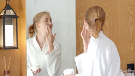 applying face cream, woman in bathrobe looking at mirror in bathroom
