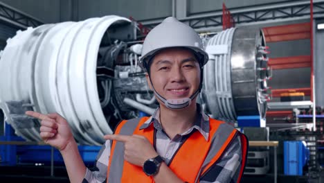 close up of asian male engineer with safety helmet smiling and pointing to side while standing with airplane engine maintenance conducted, analytics checking the turbine