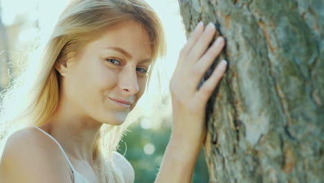 portrait of a beautiful young woman standing by the tree in the sun smiling looking at the camera co