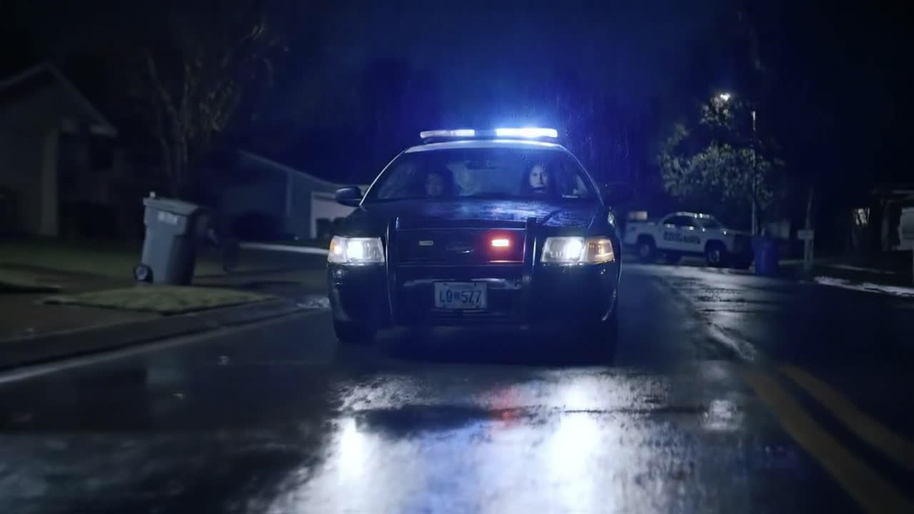 A police car with flashing lights is parked on a rainy street, highlighting the quiet neighborhood atmosphere. Officers are present, creating a tense yet controlled environment at night.