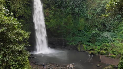 a drone flies closer to the nungnung waterfall in bali, indonesia, as a couple contemplates a swim in the pool below the falls