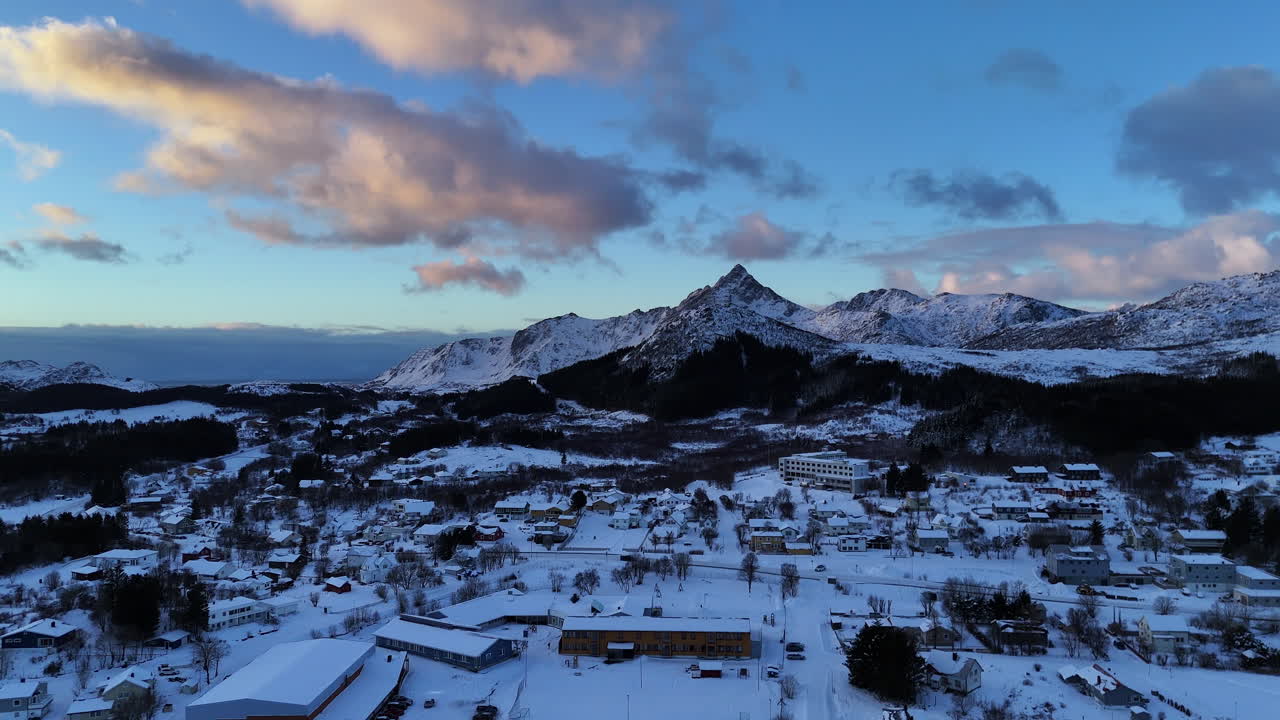 Cinematic aerial view of snow-covered mountains in Lofoten, Norway, dramatic winter landscape with icy valley, scattered houses and evening light over rugged arctic peaks