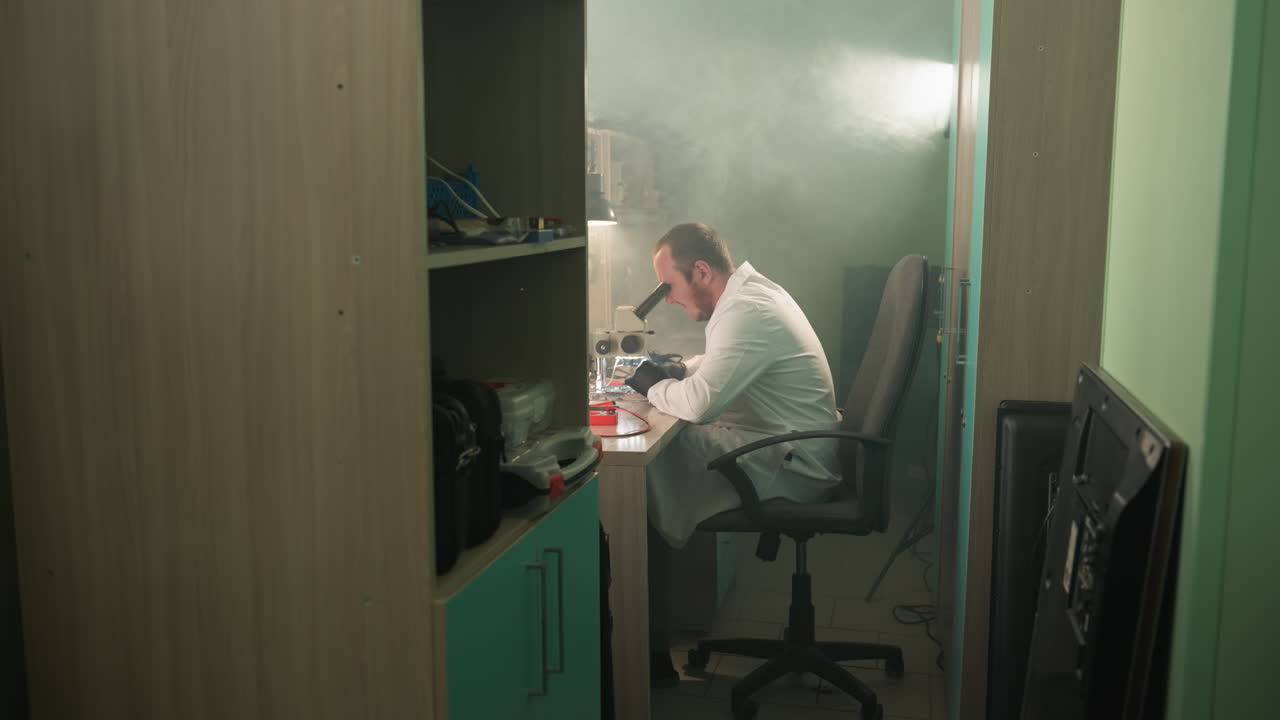 A close view of a technician in a lab coat working on an electronic components using a microscope , with a shelf by the side having other tools in it
