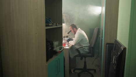 a close view of a technician in a lab coat working on an electronic components using a microscope , with a shelf by the side having other tools in it