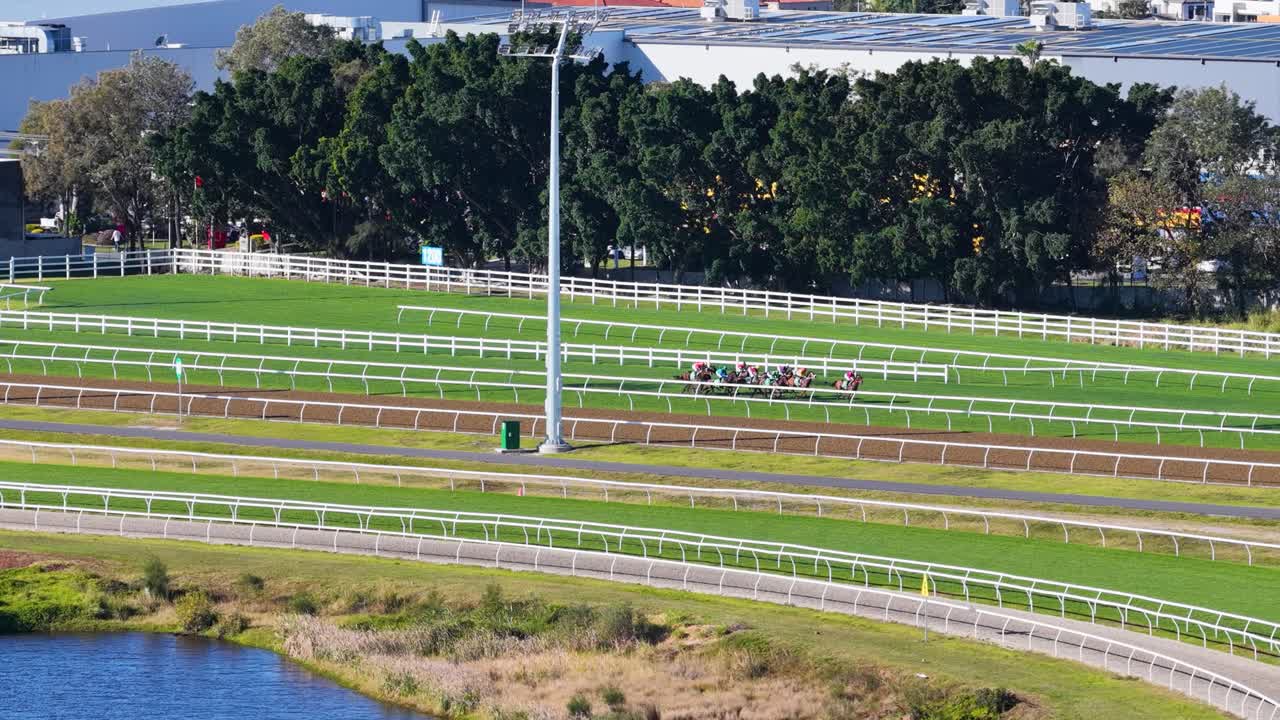 Jockeys ride thoroughbred horses at speed around a sunlit, green racetrack in Gold Coast, Australia. Wide, steady camera captures outdoor sporting event