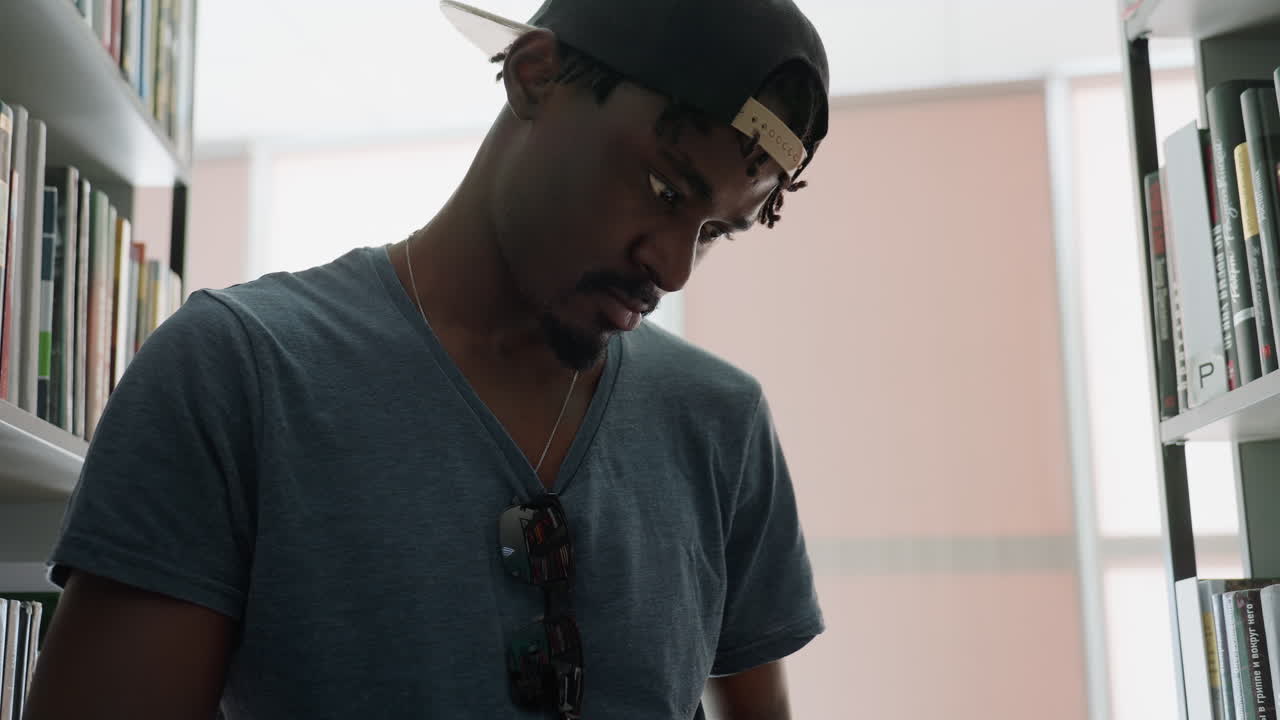 primer plano de un estudiante con gorra y gafas de sol en la camisa leyendo un libro pensativamente entre las estanterías de la biblioteca, los dedos concentrados pasan las páginas bajo la suave luz de la ventana, ambiente de sala de estudio