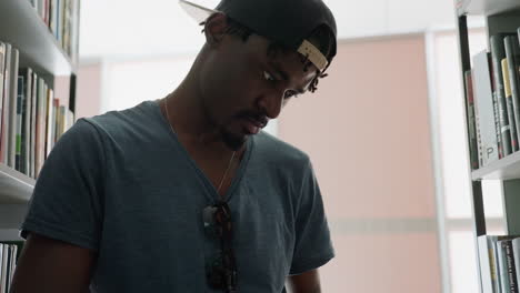 close up student in cap with sunglasses on shirt reads book thoughtfully between library shelves, focused fingers turn pages under soft window light, study room mood