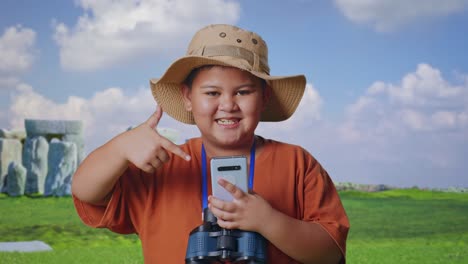 asian boy with a hat and binoculars smiling and pointing to smartphone while traveling in stonehenge. boy researcher examines something, travel tourism adventure concept, close up