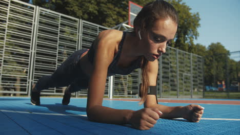 Chica-De-Fitness-Enfocada-Con-Airpods-Haciendo-Ejercicio-De-Tabla-Y-Respirando-Profundamente-En-La-Cancha-Deportiva-El-Día-De-Verano