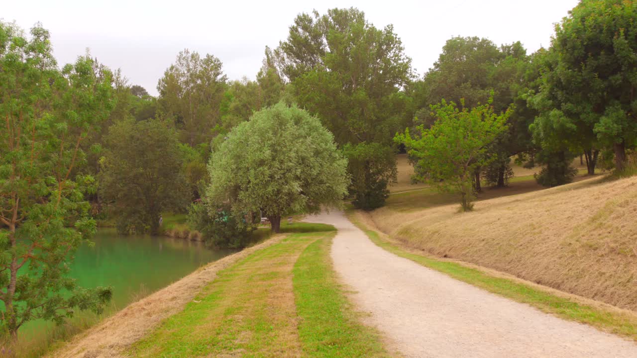 A wide shot of a dirt path running along the edge of Lac de l'Orme Blanc, flanked by trees and gentle grassy slopes