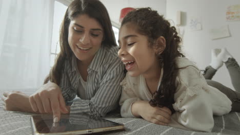 little girl and mother playing on tablet on bed