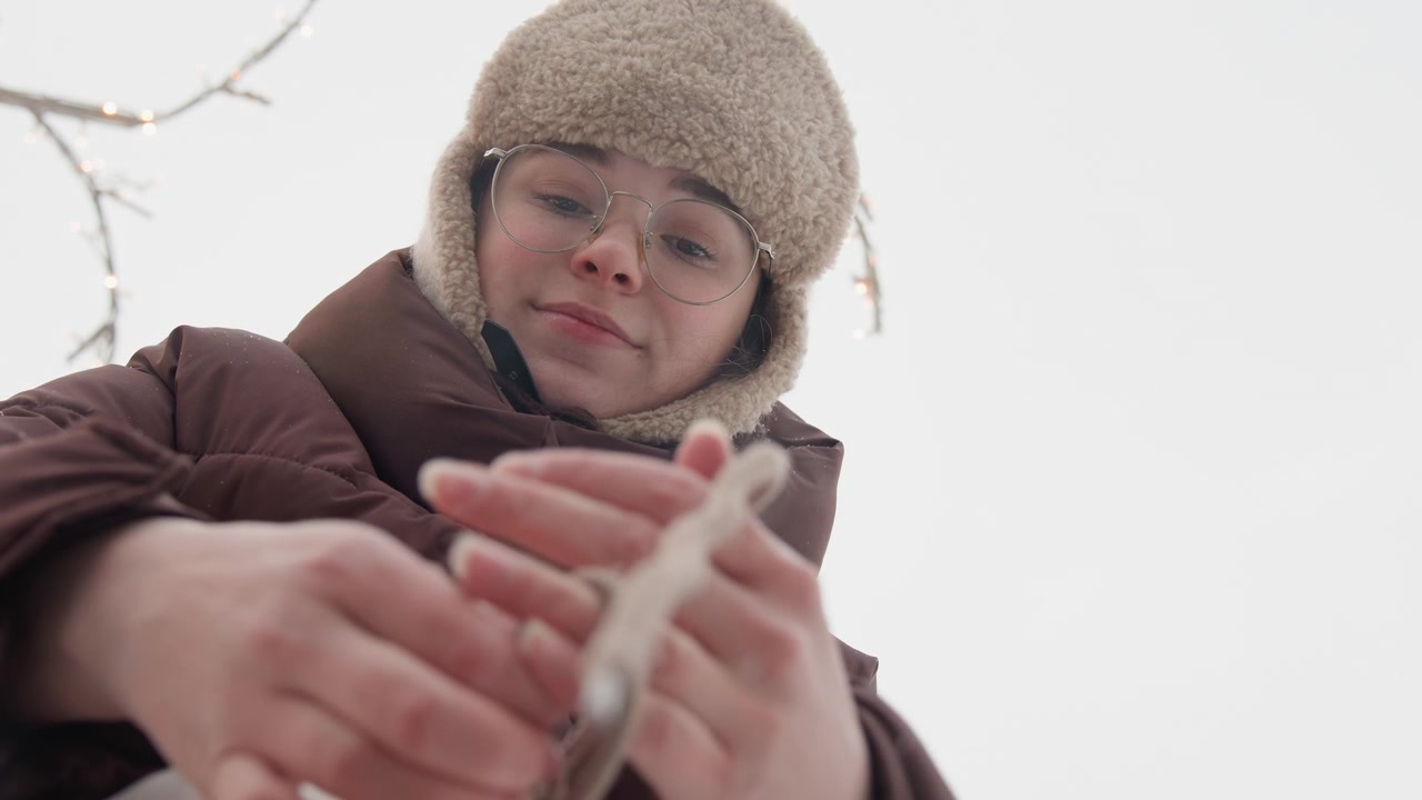 Giovane donna in un parco innevato all'aperto si china in avanti e si sistema il guanto con entrambe le mani, indossando strati invernali caldi e un cappello di lana, con alberi spogli e una luce soffusa e nuvolosa intorno a lei.