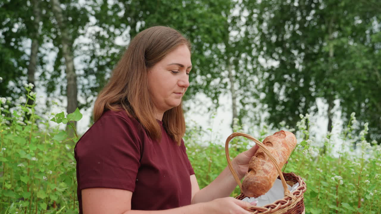 초록색 들판에서 갓 구운 빵을 바구니에 담고 빵 껍질과 텍스처를 확인하는 백인 여성, 홈 베이커 분위기, 차분한 표정, 자작나무 배경과 부드러운 여름 햇살, 일상적인 요리 의식
