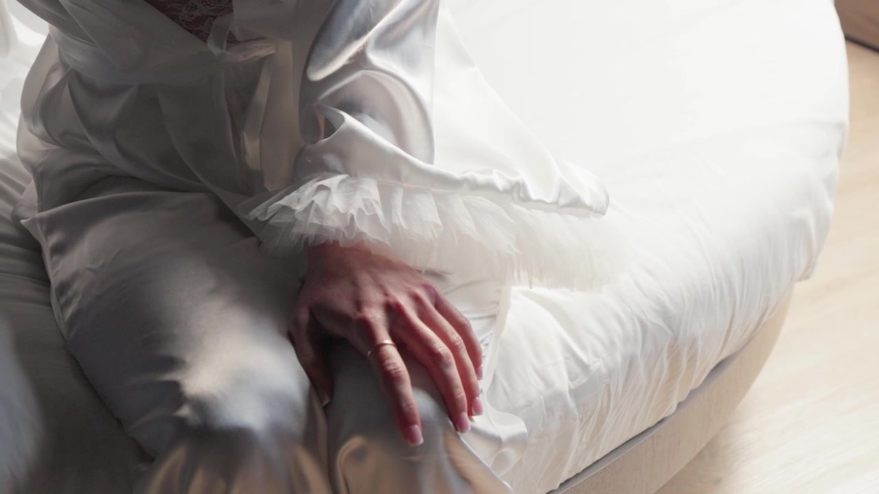 Eine detaillierte Nahaufnahme einer Braut in einem eleganten weißen Satinkleid, die auf einem hellen Bett sitzt, ihre Hand ruht anmutig auf ihrem Schoß. Natürliches, weiches Licht schafft eine ruhige, intime Stimmung