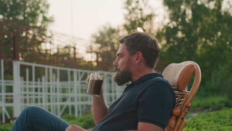 young man seated in wicker rocking chair in lush garden at sunset sipping steaming coffee from ceramic mug while unwinding in tranquil rustic outdoor setting