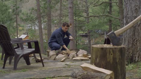 a man squats by a fire pit in the woods, carefully placing a log on the fire