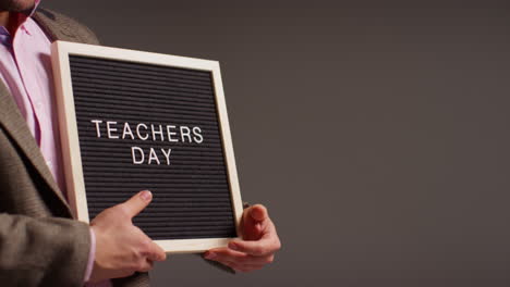 Close-Up-Studio-Shot-Of-Male-Teacher-Standing-Against-Grey-Background-Holding-Notice-Board-Reading-Teachers-Day