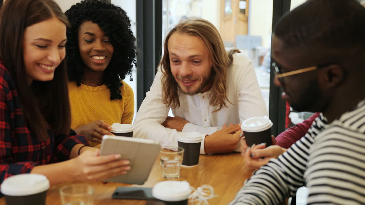 Group multiethnic group of happy friends laughing and watching a video on a tablet sitting at a table in a cafe