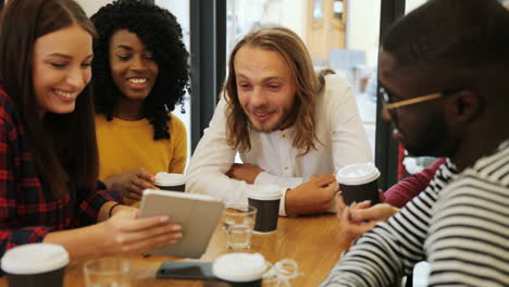 grupo multiétnico de amigos felices riéndose y viendo un video en una tableta sentados en una mesa en un café