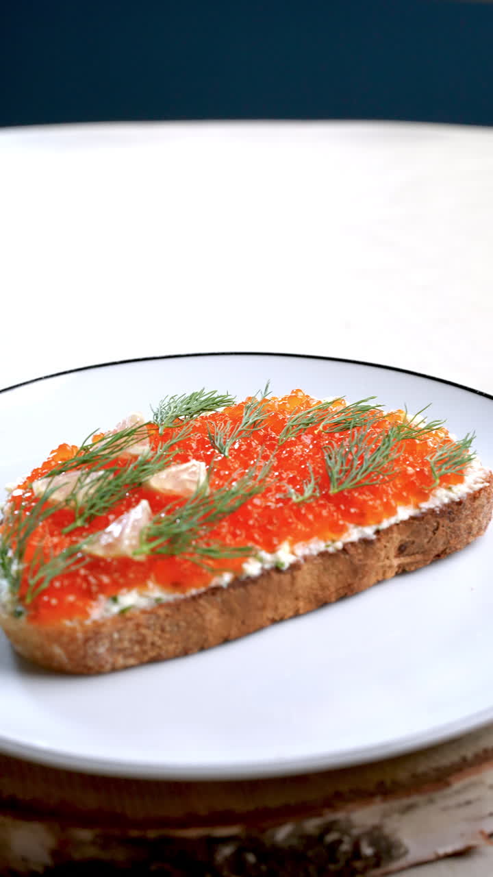 Identical salmon roe canapes, garnished with dill and lemon, rotate slightly on a white plate with a dark rim, placed on a birch wood slice against a white background and a dark blue backdrop