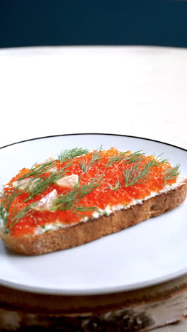 identical salmon roe canapes, garnished with dill and lemon, rotate slightly on a white plate with a dark rim, placed on a birch wood slice against a white background and a dark blue backdrop