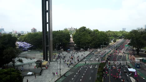 Disparo-Rápido-De-Un-Dron-Hacia-Atrás-De-Los-Corredores-De-Maratón-De-La-Ciudad-Cuando-Pasan-Por-El-Monumento-Llamado-Rastro-De-Luz