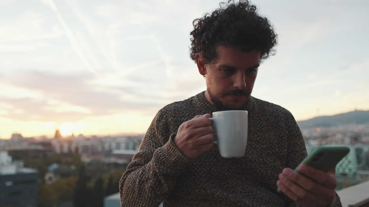 Man drinking coffee while using phone on rooftop at sunset