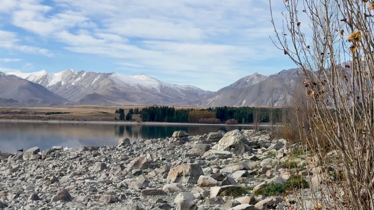 A serene view of Lake Tekapo with snow-capped mountains and rocky shoreline under clear skies in Canterbury, New Zealand