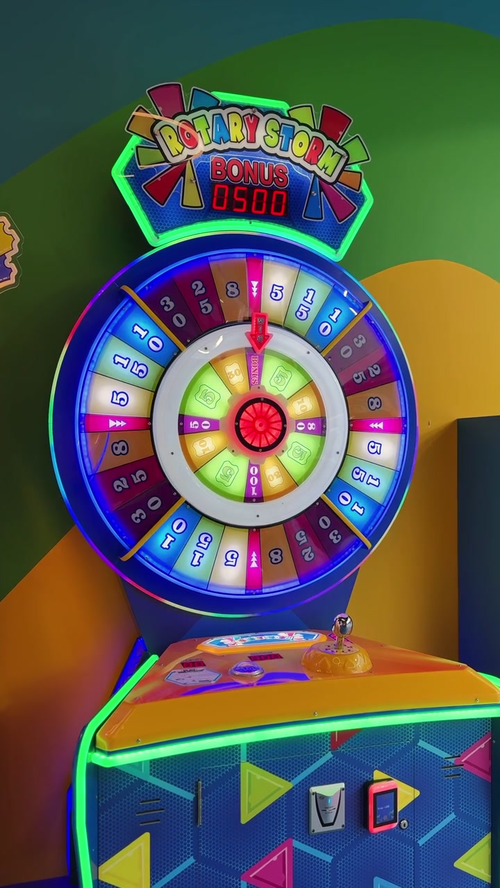Una luminosa e colorata macchina da gioco arcade con ruota della fortuna risplende all'interno di un centro divertimenti al coperto illuminato al neon. Il gioco a premi presenta segmenti premio numerati e un display digitale del punteggio bonus