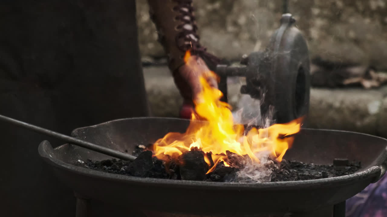 Static close-up of a blacksmith turning the hand crank of a coal forge to fan the flames and heat a metal rod; smoke, embers, and warm orange glow